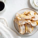 tavolo bianco da colazione con chiacchiere senza glutine in un piatto bianco, sulla sinistra una tazza di caffè, accanto un tovagliolo bianco