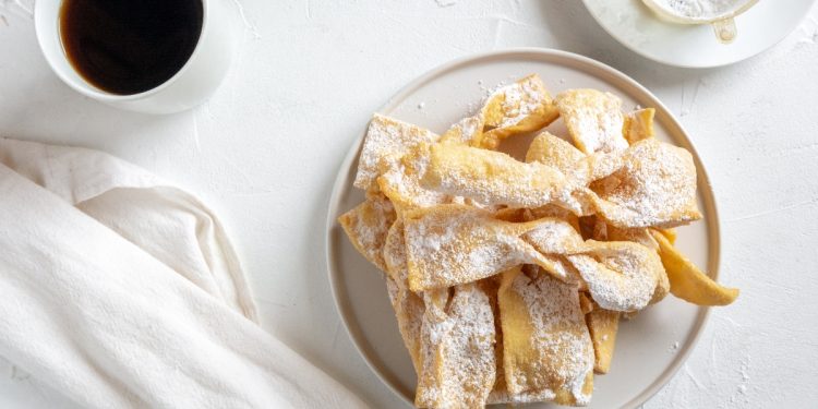 tavolo bianco da colazione con chiacchiere senza glutine in un piatto bianco, sulla sinistra una tazza di caffè, accanto un tovagliolo bianco