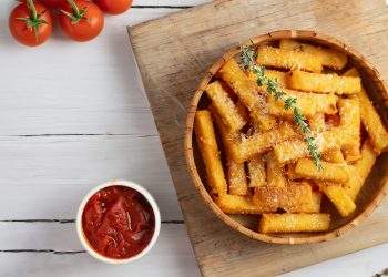 su tavolo bianco tagliere di legno con ciotola con la polenta al forno e parmigiano grattugiato e erbe, accanto piccola ciotola con salsa di pomodoro e dietro pomodorini