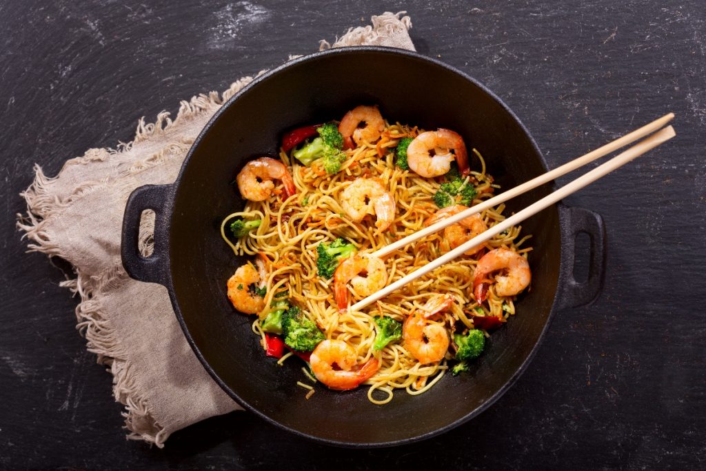 su tavolo scuro una padella wok con i noodles con gamberi e verdure e delle bacchette cinese . Sotto il wok canovaccio beige