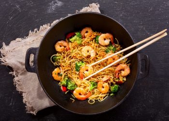 su tavolo scuro una padella wok con i noodles con gamberi e verdure e delle bacchette cinese . Sotto il wok canovaccio beige