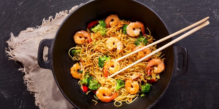 su tavolo scuro una padella wok con i noodles con gamberi e verdure e delle bacchette cinese . Sotto il wok canovaccio beige